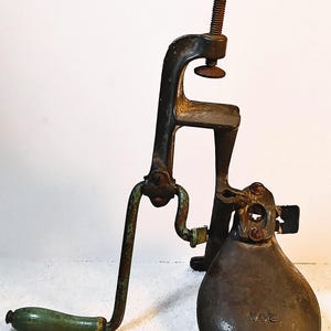 May include: Antique cast iron meat grinder with a green wooden handle and a clamp. The grinder has a dark, aged appearance with visible wear and tear, suggesting its vintage. The base has the text "V03".