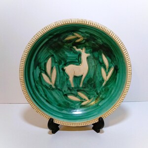 May include: A round ceramic plate featuring a deep green glazed interior with a central cream-colored deer silhouette surrounded by four cream-colored leaf motifs. The plate has a textured, cream-colored rim with vertical etched lines.