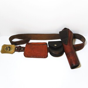 May include: A brown leather belt featuring a gold-toned rectangular buckle with a horse head relief. The belt includes a rectangular pouch, a small circular snap-closure pouch, and a long holster, all crafted from brown leather with stitching.