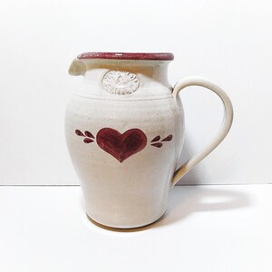 May include: A cream-colored ceramic pitcher featuring a dark red painted heart on the front, a matching red rim, and a circular embossed stamp near the spout. The pitcher has a curved handle on the right side and a smooth, glossy finish throughout.