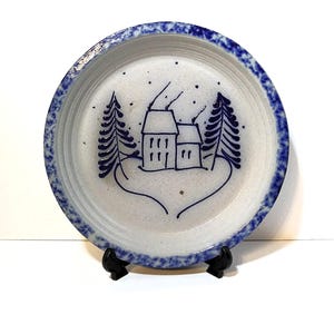 May include: A round ceramic plate with a speckled light gray base and a dark blue sponge-painted rim. The center features a hand-painted dark blue illustration of a house with two chimneys and smoke, flanked by two pine trees on a snowy ground.