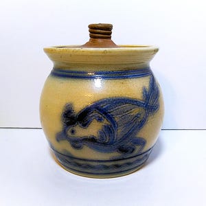 May include: A round, tan stoneware crock with a matching lid featuring a brown knob. The body is decorated with a blue hand-painted bird motif and horizontal blue bands, including a wavy line pattern near the base of the vessel.