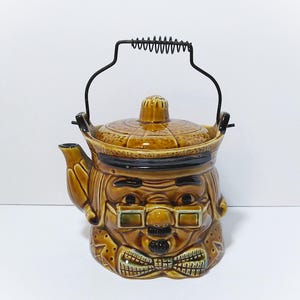 May include: A brown ceramic teapot shaped like a person's head wearing glasses and a bow tie. The teapot features a textured lid, a dark brown spout, and a black metal wire handle with a coiled spring grip at the top. The finish is glossy.