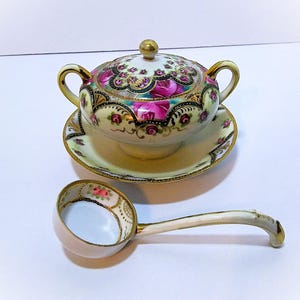 May include: A porcelain sugar bowl with a matching lid and saucer, featuring a cream-colored base, vibrant pink rose patterns, and intricate gold trim. A matching porcelain ladle with a floral interior and gold-gilded edges is included.