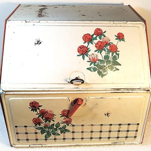 May include: Vintage metal bread box with a hinged lid. The box is painted white and cream with red trim. It features a floral design of red clover blossoms and green leaves, along with a plaid pattern and a bee.