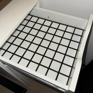 May include: An open white drawer featuring a black and white chequered pattern on the base. The drawer is part of a white cabinet or desk, with a section of the cabinet visible.
