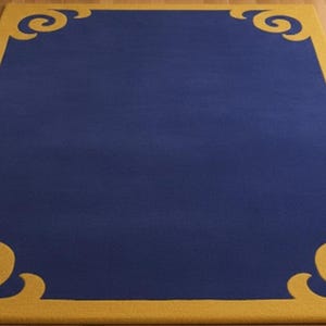 Puede incluir: Una alfombra rectangular con un centro azul marino y un borde dorado. El borde dorado presenta adornos decorativos en cada esquina. La alfombra está colocada sobre un suelo de madera clara.