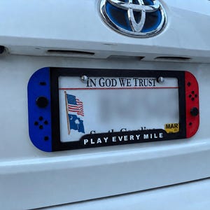 May include: A black license plate frame featuring a blue controller-style side on the left and a red controller-style side on the right. The bottom of the frame displays the white text PLAY EVERY MILE. It is mounted on a white vehicle.