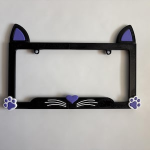 May include: A black rectangular license plate frame featuring a cat-themed design. The top corners have purple cat ears, while the bottom edge displays two white and purple paw prints, a central purple heart, and white whiskers on each side.
