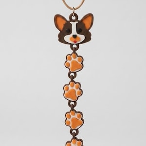 May include: A vertical pendant necklace featuring a brown and white dog face at the top, followed by four orange and white paw print charms connected by small brown rings, ending with a single white bone-shaped charm on a thin tan cord.