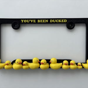 May include: A black rectangular license plate frame featuring the yellow text "YOU'VE BEEN DUCKED" at the top. The bottom edge of the frame is decorated with a row of eleven small, yellow rubber ducks of varying sizes, all facing forward.