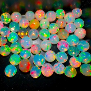 以下が含まれることがあります： 乳白色のベースにネオングリーン、鮮やかなオレンジ、エレクトリックブルー、ホットピンクの鮮やかな輝きを放つ、丸い半透明のオパールカボションのコレクション。無地の黒い表面に配置されています。