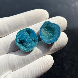 May include: Two halves of a geode, each with a vibrant turquoise interior and a rough, textured exterior. The geode halves are held against a white glove, showcasing the bright blue crystals within. The background is dark.