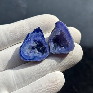 May include: Two blue geode halves with crystal formations inside. The geodes have a dark blue outer edge and a lighter blue interior. The geodes are held in a white gloved hand against a dark background. The geodes are a natural, decorative item.