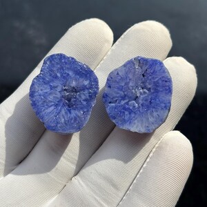 Könnte beinhalten: Zwei polierte, blau-violette Achatsgeoden, jede mit einem kristallinen Inneren. Die Geoden werden in einer weißen behandschuhten Hand gehalten, die ihre komplizierten, natürlichen Formationen zeigt. Die Farbe geht von einem tiefen Blau in einen helleren, fast weißen Farbton über.