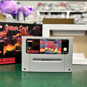 May include: A Super Nintendo video game cartridge and box set. The cartridge is light grey with a colourful label featuring a winged demon. The box is black with the game title "Demon's Crest" and a fiery illustration. A white frame is also present.