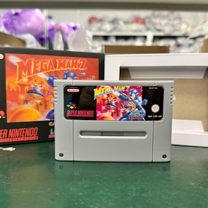 Puede incluir: Un cartucho y una caja de videojuego Super Nintendo para Mega Man 7. El cartucho es gris con una etiqueta colorida con Mega Man. La caja es negra con el título del juego en naranja y una ilustración vibrante. Una caja blanca está abierta junto al juego.