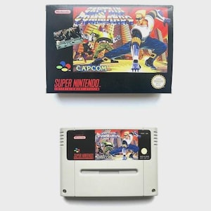 May include: A Super Nintendo video game cartridge and box set for "Captain Commando." The box is black with colourful graphics of the game's characters. The cartridge is light grey with a matching label.