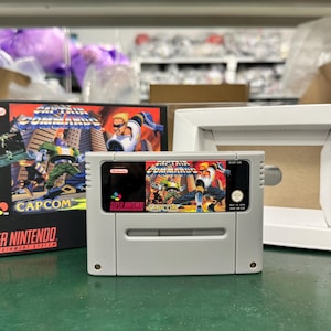 Op de afbeelding: Een Super Nintendo-videogamecartridge en -doos voor "Captain Commando" van Capcom. De cartridge is grijs met een kleurrijk label. De doos bevat actievolle illustraties met de titel en het logo van de game. Een witte fotolijst staat ook in de afbeelding.