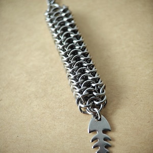 May include: A silver chainmail bracelet with a fishbone charm. The bracelet is made of small, interlocking metal rings. The fishbone charm is a simple, stylized design.