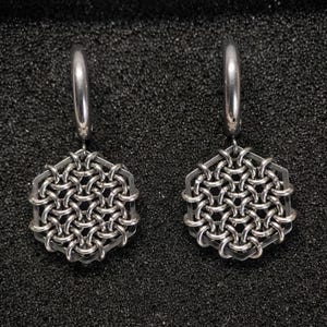Könnte beinhalten: Ein Paar silberfarbene Ohrringe. Jeder Ohrring hat einen sechseckigen Rahmen mit einem gewebten Kettenmuster. Die Ohrringe haben einen Creolenverschluss. Der Hintergrund ist eine strukturierte schwarze Oberfläche.
