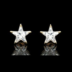 以下が含まれることがあります： 星型のスタッドピアス。各イヤリングは、金色の金属にセットされた透明でファセットカットされた宝石が特徴です。イヤリングは黒い背景に表示され、その下の反射が見えます。