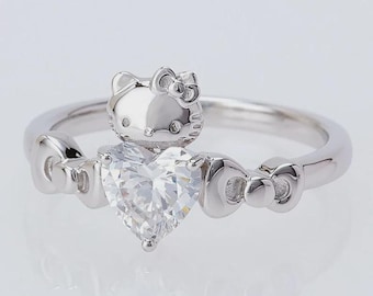 May include: A silver ring featuring a heart-shaped clear gemstone. The ring has a Hello Kitty design on top of the gemstone and bow accents on the sides. The ring is set against a white background.