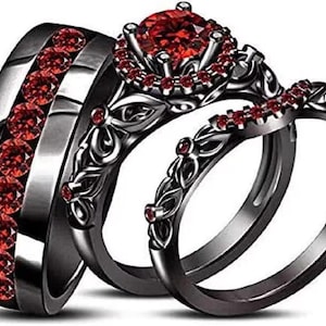 May include: A three-piece black wedding ring set. The set includes a wide band with red gemstones, a floral design ring with a large red centre stone, and a matching band with red accents. The rings have a dark, metallic finish.