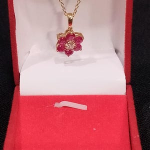 Könnte beinhalten: Eine goldfarbene Halskette mit einem blütenförmigen Anhänger, besetzt mit roten Edelsteinen und einem zentralen klaren Stein. Die Kette wird in einer offenen roten Schmuckschatulle mit weißem Innenraum vor dunklem Hintergrund präsentiert.