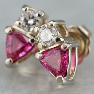 May include: Close-up of a pair of stud earrings. Each earring features a triangular pink gemstone and a round, clear gemstone set in a silver-coloured metal. The earrings have a classic, elegant design.
