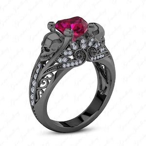 Puede incluir: Anillo negro con calaveras y una gran gema roja facetada. El anillo presenta dos calaveras a cada lado de la gema, con detalles florales y de volutas. Pequeñas gemas transparentes están engastadas a lo largo de la banda y en los detalles florales.