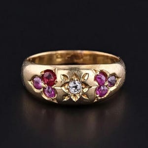 Op de afbeelding: Een gouden ring met een centrale diamant en robijnaccenten. De ring heeft een bloemenontwerp met een centrale diamant en clusters van robijnen en roze edelstenen aan weerszijden. De ring staat tegen een zwarte achtergrond.