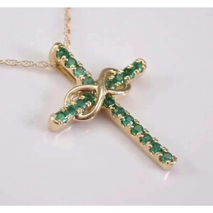 May include: A gold-colored pendant necklace featuring a cross design. The cross is adorned with green gemstones and has a unique knot detail at the center. The necklace is displayed on a white surface.