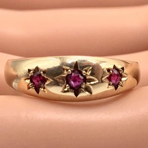 Könnte beinhalten: Ein goldener Ring mit drei sternförmigen Designs, die jeweils einen leuchtend roten Edelstein halten. Der Ring hat ein glattes, poliertes Band und einen klassischen, eleganten Stil. Die Edelsteine sind in einer Krappenfassung gefasst.