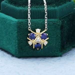 May include: A silver chain necklace with a pendant featuring a gold-colored geometric design. The pendant has three dark blue gemstones, three small white gemstones, and a white triangular gemstone in the center. The necklace is displayed on a dark green velvet surface.