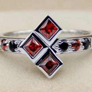 May include: Silver ring featuring a geometric design with three square, red gemstones and black gemstones. The ring has a polished finish and is set with small, round black and red stones along the band.