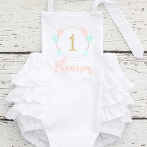 May include: White romper with a textured fabric and ruffled trim. The romper features an embroidered design with the number 1, balloons, and the name "Eleanor" in a cursive font. The straps are white and tie at the shoulders.