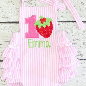 May include: Pink and white striped baby romper with ruffled sleeves. Features a large number 1, a strawberry applique, and the name "Emma" embroidered in green. The romper has tie straps.