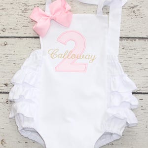 May include: White baby romper with pink number "2" and the name "Calloway" embroidered in gold. The romper features a pink bow and white ruffles. The straps are white.