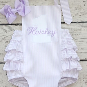May include: A light purple and white pinstriped romper featuring a large white number one with the name Haisley embroidered in purple script across the center. The garment includes ruffled sides and a matching light purple hair bow accessory.