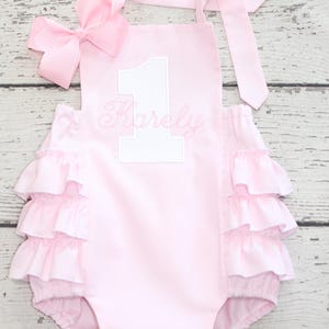 May include: A light pink romper featuring three tiers of ruffles on each side, a large pink bow attached to the shoulder strap, and a white applique number one with the name Karely embroidered in pink script lettering on the front chest area.