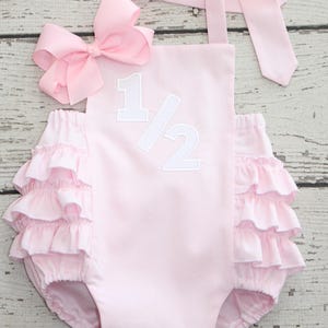 May include: A light pink baby romper with the number "1/2" in white. The romper has ruffled trim around the legs and a large pink bow. The straps tie at the shoulders.