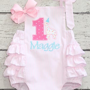 May include: A light pink and white pinstriped romper featuring a large pink polka-dot number one, a giraffe wearing a party hat, and the name Maggie embroidered in blue. The garment includes a pink hair bow and two tiers of ruffles on each side.