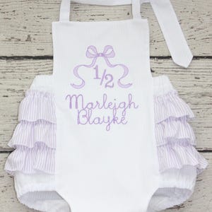 May include: White baby romper with lavender accents. The romper features a bow and the text "1/2 Marleigh Blayke" embroidered in lavender. The bottom has ruffled lavender and white striped fabric.