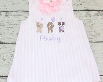 Puede incluir: Un vestido sin mangas de rayas finas en color rosa claro y blanco con un lazo grande rosa en el cuello, dos botones en los hombros y un bordado de tres perros con gorros de fiesta y un globo sobre el nombre bordado Paisley.