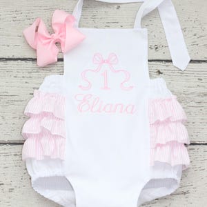 May include: A white romper with pink striped ruffles on the sides, featuring an embroidered pink bow, the number 1, and the name Eliana in pink thread. A matching pink hair bow is included with the white romper, which has tie-up shoulder straps.