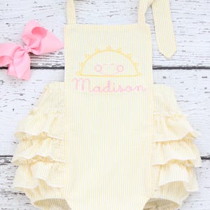 May include: Yellow and white striped romper with a sun design and the name "Madison" embroidered in pink. The romper has ruffled trim and a matching pink bow.