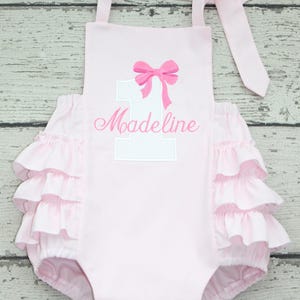 May include: A light pink romper featuring a white number one with a pink bow graphic and the name Madeline embroidered in pink script. The garment has ruffled sides and tie-top shoulder straps, providing a decorative look for a first birthday.