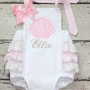 May include: A white romper featuring a pink embroidered seashell and the name Ellie in gold script. The garment has pink and white striped ruffled sides, a matching striped tie strap, and a large pink hair bow attached to the top left corner.