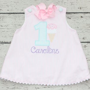 May include: Pink and white striped dress with a large pink bow at the top. The dress features a number 1 and an ice cream cone applique, with the name "Caroline" embroidered below. The dress has a scalloped hem.